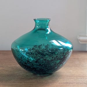 Puede incluir: Un jarr&oacute;n de vidrio color teal con cuerpo redondeado y cuello estrecho. La parte inferior del jarr&oacute;n presenta un patr&oacute;n moteado, teal oscuro, creando una apariencia texturizada. El jarr&oacute;n est&aacute; sobre una superficie de madera.