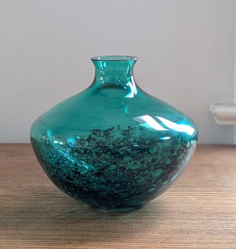 Puede incluir: Un jarr&oacute;n de vidrio color teal con cuerpo redondeado y cuello estrecho. La parte inferior del jarr&oacute;n presenta un patr&oacute;n moteado, teal oscuro, creando una apariencia texturizada. El jarr&oacute;n est&aacute; sobre una superficie de madera.