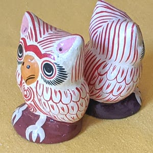 May include: Two small owl figurines featuring a white base with intricate red line patterns. Each owl has a yellow beak, black eyes with long lashes, and pink ear tufts. The owls are perched on dark brown circular bases, showcasing detailed hand-painted designs.