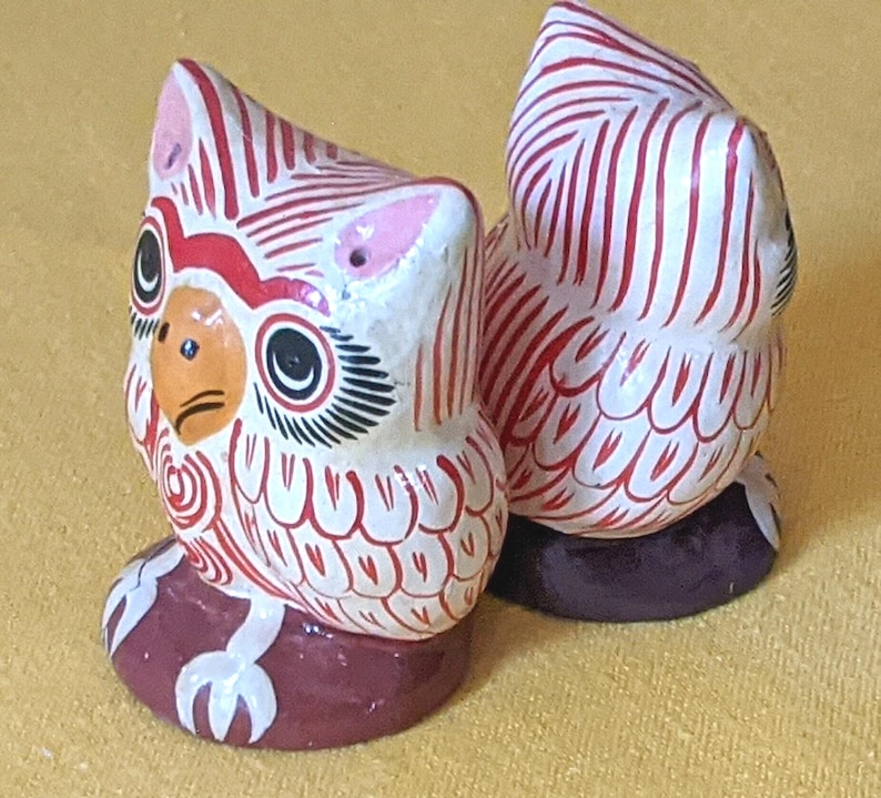 May include: Two small owl figurines featuring a white base with intricate red line patterns. Each owl has a yellow beak, black eyes with long lashes, and pink ear tufts. The owls are perched on dark brown circular bases, showcasing detailed hand-painted designs.