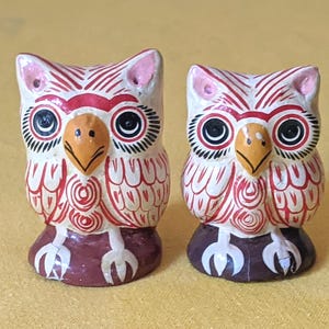 May include: Two small owl figurines featuring white bodies with intricate red line patterns, yellow beaks, and large black eyes. Each owl has pink-tipped ears and stands on a dark brown base with white claw details, showcasing a hand-painted aesthetic.