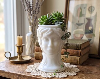 May include: A white milk glass head-shaped planter featuring a sculpted face with wavy hair details. The planter contains a green succulent and a trailing string of hearts plant, resting on a round cream-colored crocheted doily.
