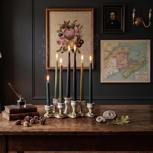 May include: A multi-tiered ceramic candelabra holding six lit taper candles in shades of green, black, and brown. The base features a textured, cream-colored finish with ornate detailing. The candles are arranged at varying heights on the table.