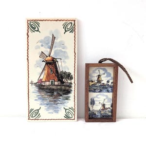 May include: A rectangular ceramic tile featuring a hand-painted windmill scene with a brown border and green corner accents, displayed alongside a smaller, framed wooden wall hanging that contains two stacked miniature windmill illustrations.