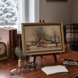 May include: A framed landscape print titled "Returning to the Farm" depicting a snowy rural scene with a red barn, bare trees, and cattle. The print is displayed on a wooden easel, accompanied by a brass clock, books, candles, and a fountain pen.