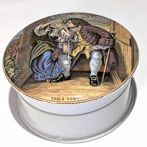 May include: A white ceramic jar with a lid depicting a detailed illustration of two figures in historical attire. The lid has a gold dotted border, and the text "UNCLE TOBY" is displayed. The jar has a clear, rounded base.