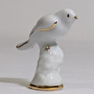 May include: A white porcelain bird figurine perched on a textured base. The bird features gold-painted accents on its beak, wing edges, and tail feathers. The base of the figurine is also trimmed with a thin, circular gold band at the bottom edge.