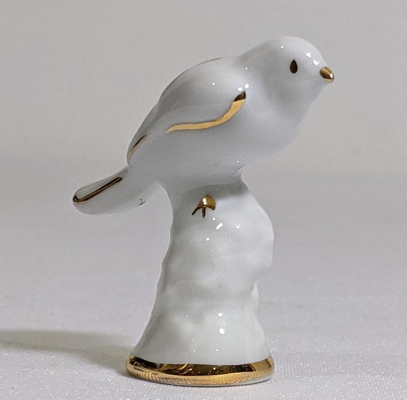 May include: A white porcelain bird figurine perched on a textured base. The bird features gold-painted accents on its beak, wing edges, and tail feathers. The base of the figurine is also trimmed with a thin, circular gold band at the bottom edge.