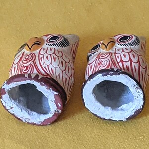May include: Two hand-painted owl figurines featuring a cream-colored base with intricate red line patterns, black and white eyes, and yellow beaks. Each owl has a hollow, circular base painted in a dark reddish-brown color with a white interior.