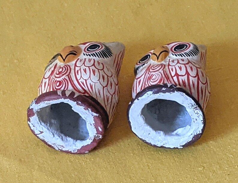 May include: Two hand-painted owl figurines featuring a cream-colored base with intricate red line patterns, black and white eyes, and yellow beaks. Each owl has a hollow, circular base painted in a dark reddish-brown color with a white interior.