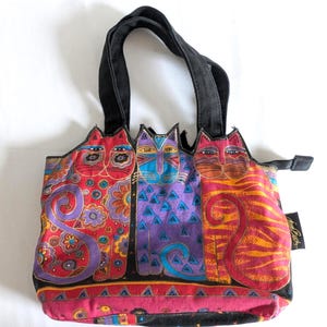 Puede incluir: Un bolso de tela con un estampado gr&aacute;fico colorido de tres gatos estilizados en rojo, morado y naranja. El bolso tiene asas negras, cierre de cremallera y una etiqueta lateral negra con texto en blanco. Los dise&ntilde;os de los gatos incluyen patrones.
