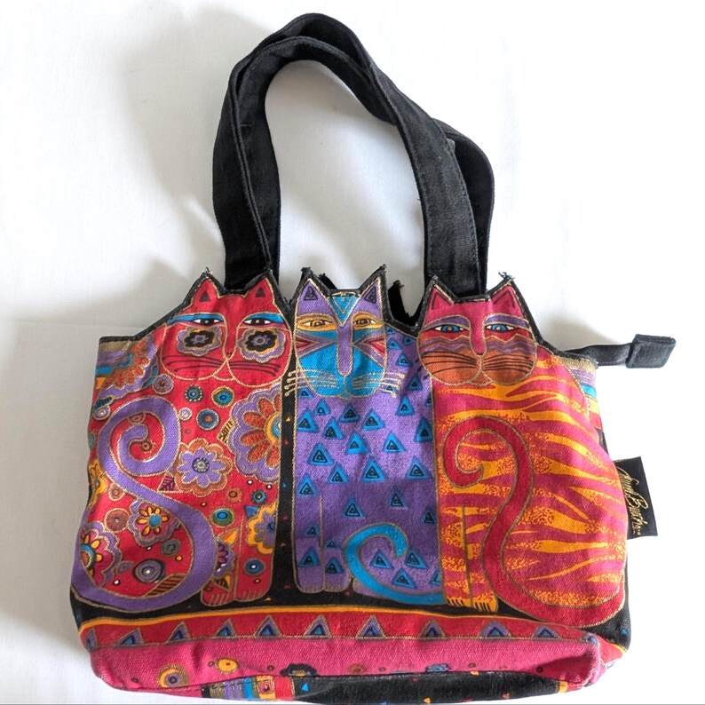 Puede incluir: Un bolso de tela con un estampado gr&aacute;fico colorido de tres gatos estilizados en rojo, morado y naranja. El bolso tiene asas negras, cierre de cremallera y una etiqueta lateral negra con texto en blanco. Los dise&ntilde;os de los gatos incluyen patrones.