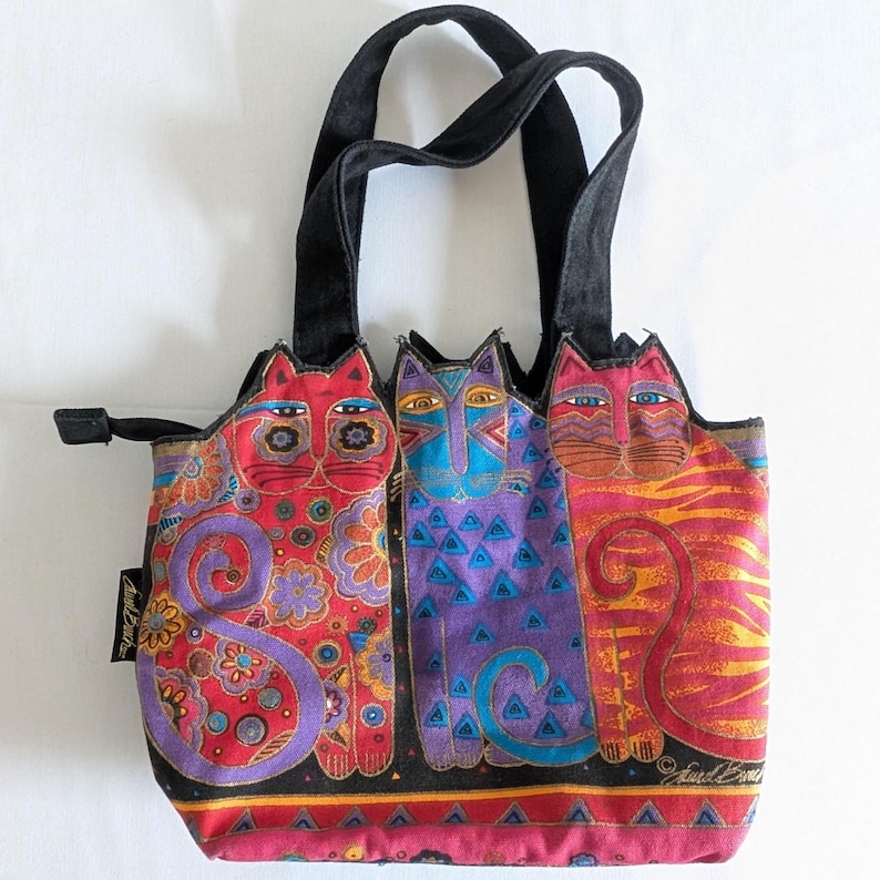 Puede incluir: Un bolso de mano con tres gatos estilizados en rojo, morado y naranja con intrincados patrones florales y geom&eacute;tricos. El bolso tiene asas de tela negra, cierre de cremallera y una etiqueta negra en el lateral con el texto Laurel Burch.