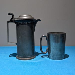 May include: A vintage pewter coffee pot with a hinged lid and a handle, next to a matching pewter mug with a curved handle. The objects are displayed on a blue surface against a gray background.