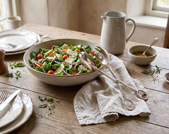 May include: A large, speckled ceramic bowl filled with a fresh salad of green leaves, red cherry tomatoes, purple onion slices, and shaved parmesan cheese, accompanied by a pair of silver-toned metal salad tongs with ring-shaped handles.