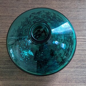 Puede incluir: Una botella de vidrio color verde azulado con base redonda y cuello cil&iacute;ndrico. El vidrio tiene una textura burbujeante y refleja la luz. La botella est&aacute; sobre una superficie de madera.