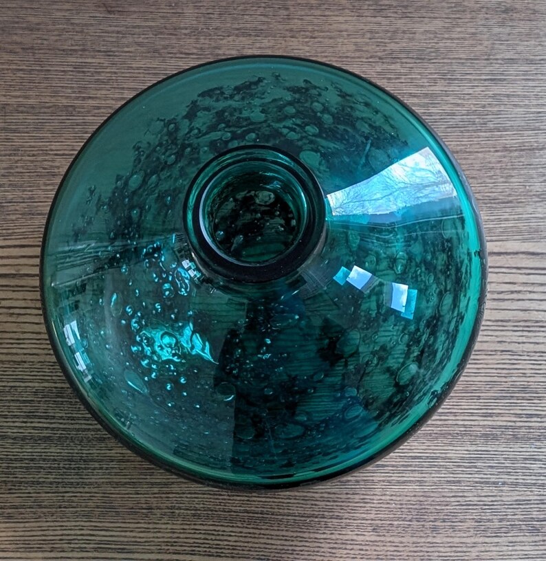 Puede incluir: Una botella de vidrio color verde azulado con base redonda y cuello cil&iacute;ndrico. El vidrio tiene una textura burbujeante y refleja la luz. La botella est&aacute; sobre una superficie de madera.