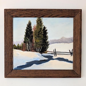 May include: Oil painting depicting a winter scene with snow, evergreen trees, and a wooden fence, set within a dark brown wooden frame. The sky is a pale blue. The painting evokes a sense of calm.