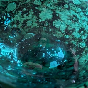 Puede incluir: Primer plano de un cuenco de vidrio de color verde azulado con una superficie texturizada. El cuenco tiene un patr&oacute;n oscuro y moteado y numerosas formaciones peque&ntilde;as en forma de burbujas. El vidrio refleja la luz, creando reflejos brillantes.