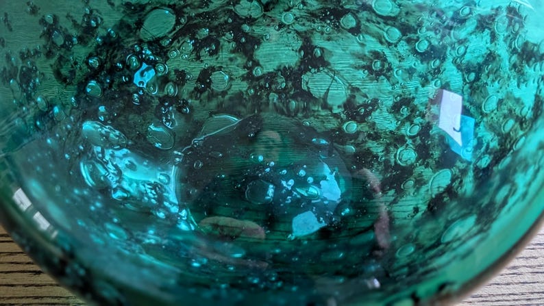 Puede incluir: Primer plano de un cuenco de vidrio de color verde azulado con una superficie texturizada. El cuenco tiene un patr&oacute;n oscuro y moteado y numerosas formaciones peque&ntilde;as en forma de burbujas. El vidrio refleja la luz, creando reflejos brillantes.