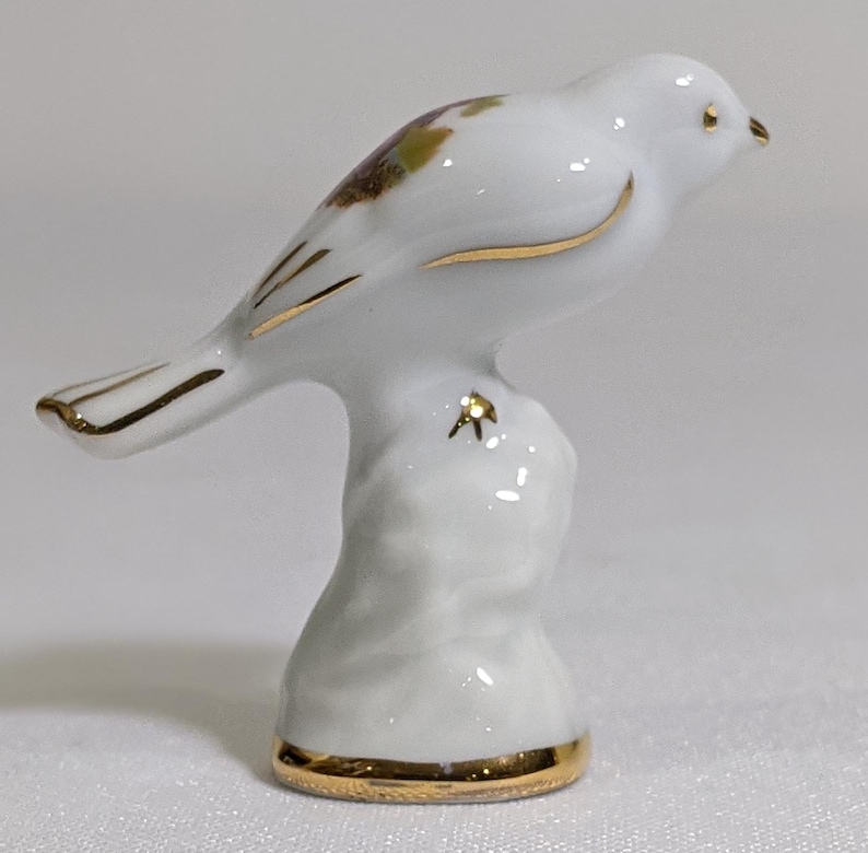 May include: A white porcelain bird figurine perched on a textured base. The bird features gold trim along its wings, tail, and beak, with a small gold accent on the base. A subtle brown and green floral pattern is visible on the bird's back.