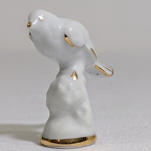 May include: A small white porcelain bird figurine perched on a textured stump. The bird features delicate gold trim on its beak, wings, and tail feathers. The base of the stump is also accented with a gold band, creating a polished finish.
