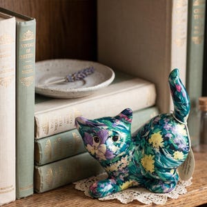 May include: A ceramic cat figurine featuring a vibrant floral pattern in shades of teal, purple, and yellow. The cat is depicted in a crouching position with its tail held upright. A small, circular paper tag is attached to the tail of the figurine.
