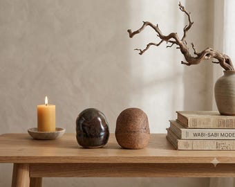 May include: A yellow pillar candle on a small wooden plate, two smooth rounded ceramic objects in dark brown and tan, a stack of three books with the text WABI-SABI MODERN on the spine, and a ceramic vase containing a twisted wooden branch.