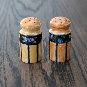 May include: A pair of vintage salt and pepper shakers. The shakers have a light orange top with multiple holes and a cylindrical body. The body is light orange with vertical black stripes and a black band with floral designs.