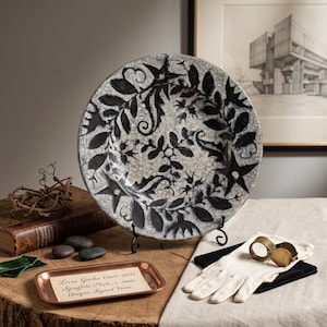 May include: A round ceramic plate featuring a black sgraffito botanical pattern on a white crackle-glazed background. The plate is displayed on a black metal stand. A small copper tray nearby contains a card with the text: Livia Gorka (1925-2011) Sgraffito Plate, c. 1960s Unique, Signed Verse.