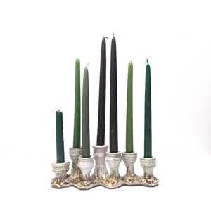 May include: A ceramic candelabra with a textured, speckled white and brown base holding six tapered candles in varying shades of green and dark grey. The candles are arranged in a staggered height pattern, with the tallest candles positioned in the centre.