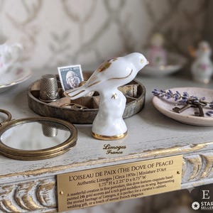 May include: A white porcelain bird figurine with gold accents, identified as L'OISEAU DE PAIX (THE DOVE OF PEACE) by Limoges France. The plaque states dimensions of 4.45 cm H x 5.72 cm D x 1.91 cm W. It features hand-painted gold details.