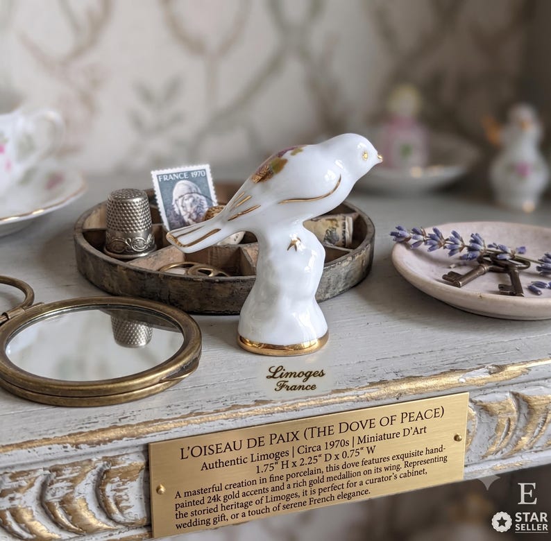 May include: A white porcelain bird figurine with gold accents, identified as L'OISEAU DE PAIX (THE DOVE OF PEACE) by Limoges France. The plaque states dimensions of 4.45 cm H x 5.72 cm D x 1.91 cm W. It features hand-painted gold details.