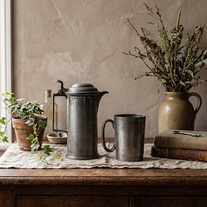 May include: A tall, cylindrical pewter lidded pitcher with a curved handle and a matching pewter mug with a simple handle. Both items feature a matte, weathered grey finish and are positioned on a light-colored fabric runner.