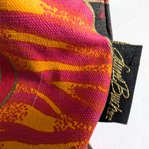 Puede incluir: Un art&iacute;culo de tela texturizada con un patr&oacute;n vibrante de rayas diagonales naranjas y rosas. Una peque&ntilde;a etiqueta rectangular negra est&aacute; unida al lateral, mostrando el texto bordado en dorado "Lori Burch" en una fuente cursiva.