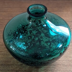 Puede incluir: Un jarr&oacute;n de vidrio de color verde azulado con un cuerpo redondo y un cuello estrecho. El vidrio tiene una apariencia texturizada y burbujeante, y el jarr&oacute;n refleja la luz, creando reflejos. El jarr&oacute;n es probablemente para fines decorativos.