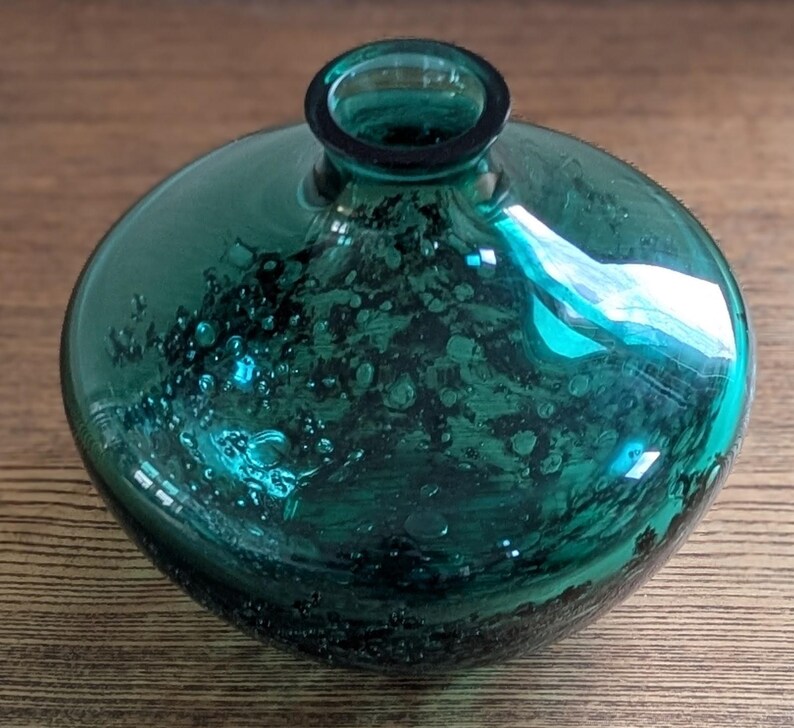 Puede incluir: Un jarr&oacute;n de vidrio de color verde azulado con un cuerpo redondo y un cuello estrecho. El vidrio tiene una apariencia texturizada y burbujeante, y el jarr&oacute;n refleja la luz, creando reflejos. El jarr&oacute;n es probablemente para fines decorativos.