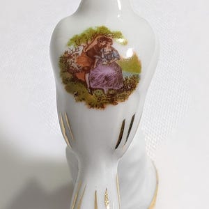 May include: A white porcelain bird figurine featuring a colourful transfer print of a couple in a landscape on its back. The bird has gold painted accents on its tail feathers and wings, with a gold rim around the base of the object.