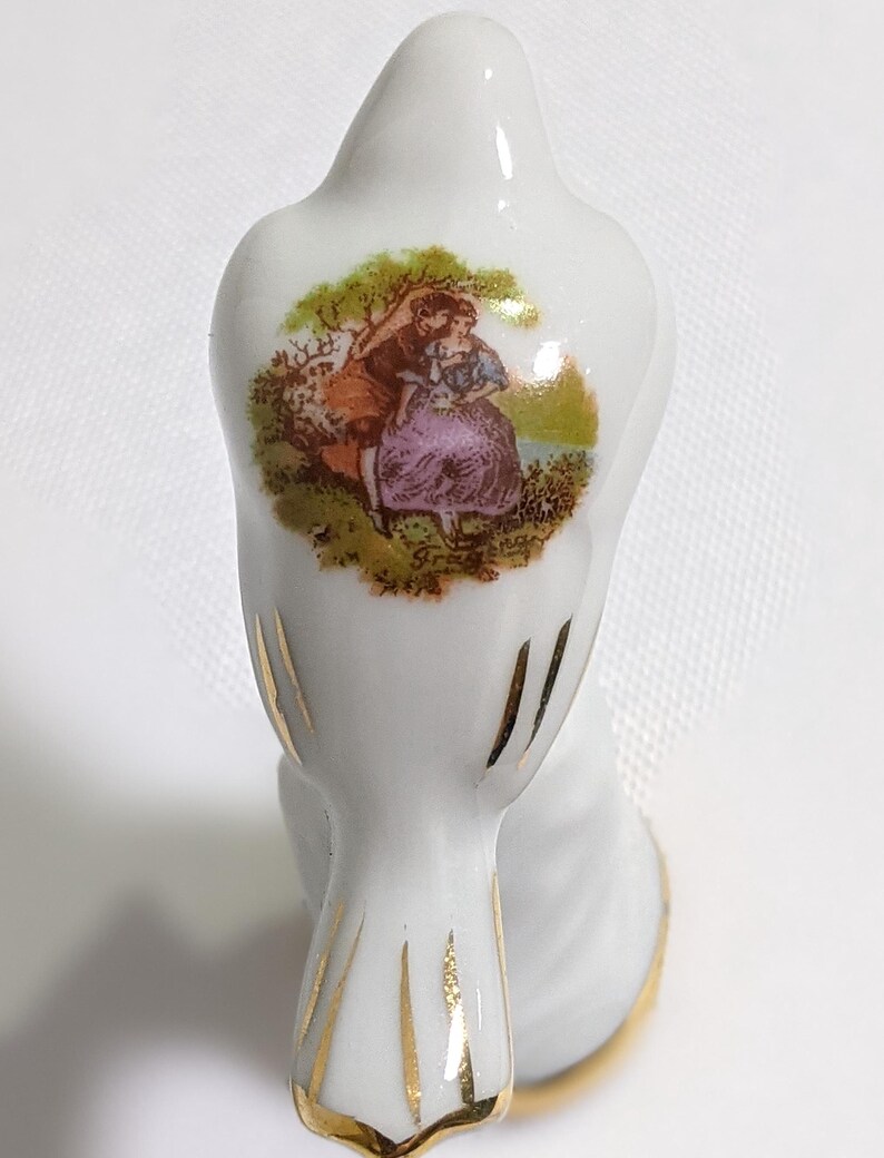 May include: A white porcelain bird figurine featuring a colourful transfer print of a couple in a landscape on its back. The bird has gold painted accents on its tail feathers and wings, with a gold rim around the base of the object.