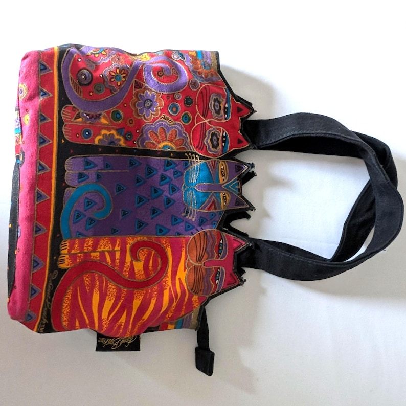 Puede incluir: Un bolso de tela con un estampado multicolor vibrante de tres gatos estilizados en tonos rojo, morado y naranja con intrincados patrones florales y geom&eacute;tricos. El bolso tiene asas de tela negra y un panel lateral negro con borde triangular.