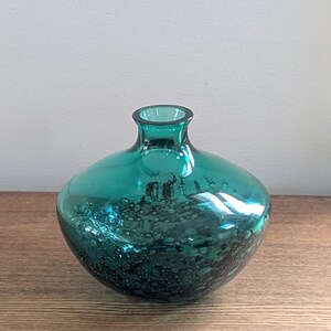 Puede incluir: Un jarr&oacute;n de vidrio color turquesa con cuerpo redondeado y cuello estrecho. El jarr&oacute;n tiene una superficie texturizada con manchas oscuras, creando un efecto visual &uacute;nico. El jarr&oacute;n est&aacute; sobre una superficie de madera.