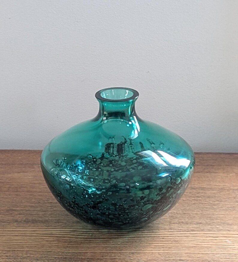 Puede incluir: Un jarr&oacute;n de vidrio color turquesa con cuerpo redondeado y cuello estrecho. El jarr&oacute;n tiene una superficie texturizada con manchas oscuras, creando un efecto visual &uacute;nico. El jarr&oacute;n est&aacute; sobre una superficie de madera.