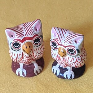 May include: Two small owl figurines featuring white bodies with red linear patterns, black and white circular eyes, yellow beaks, and pink ear tufts. Each owl stands on a dark brown circular base with white claw details painted on the front.