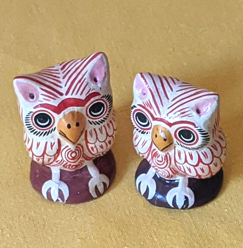 May include: Two small owl figurines featuring white bodies with red linear patterns, black and white circular eyes, yellow beaks, and pink ear tufts. Each owl stands on a dark brown circular base with white claw details painted on the front.