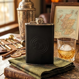 May include: A black rectangular metal flask with a circular embossed monogram featuring the letters CM. The flask has a metallic screw-top cap and is positioned on a folded green tweed fabric resting atop a book titled Whisky Legends & Lore.