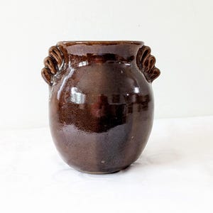 May include: A brown ceramic vase with a glossy finish. The vase has a rounded body and two decorative handles. The vase is approximately 15 cm tall and 13 cm wide.