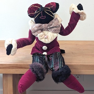 May include: A plush doll with a black face, burgundy velvet outfit, and patterned pants. The doll has a large bow tie, white trim, and faux fur accents. It is seated with arms outstretched.