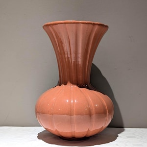 Peut inclure: Un vase en céramique pêche avec une base bulbeuse et un col évasé cannelé. Le vase présente des arêtes verticales et une finition brillante. Le vase est posé sur une surface blanche.