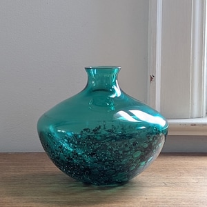 Puede incluir: Un jarr&oacute;n de vidrio color turquesa con un cuerpo redondeado y un cuello estrecho. La parte inferior del jarr&oacute;n presenta un dise&ntilde;o texturizado con burbujas. El jarr&oacute;n est&aacute; sobre una superficie de madera, con una ventana visible al fondo.