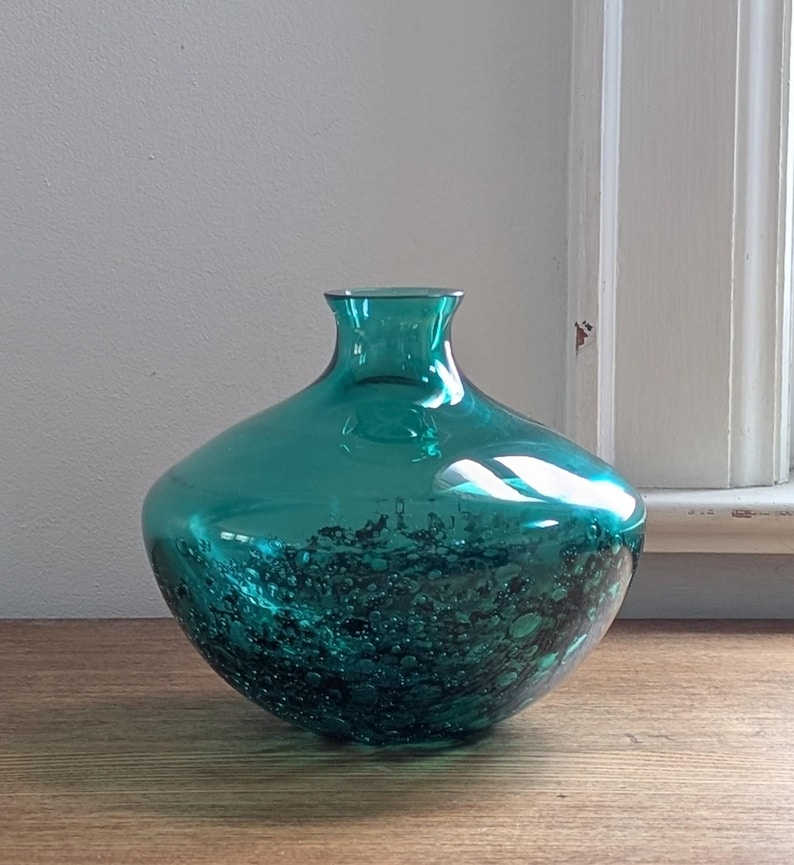 Puede incluir: Un jarr&oacute;n de vidrio color turquesa con un cuerpo redondeado y un cuello estrecho. La parte inferior del jarr&oacute;n presenta un dise&ntilde;o texturizado con burbujas. El jarr&oacute;n est&aacute; sobre una superficie de madera, con una ventana visible al fondo.